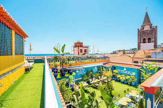 Kattoterassi, jossa on vihreitä kasveja, istumapaikkoja ja värikkäitä rakennuksia taustalla. (Tekoälyn luoma teksti)