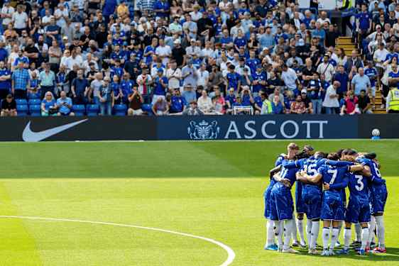Pelaajat sinisissä jalkapallopaidoissa seisovat ryhmässä ympyrässä vihreällä nurmella, taustalla yleisöä. (Tekoälyn luoma teksti)