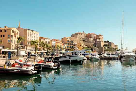 Pieni venesatama, jossa veneitä kiinnitettynä laiturille. Ympärillä värikkäitä rakennuksia ja taustalla historiallinen linna korkeudella. (Tekoälyn luoma teksti)