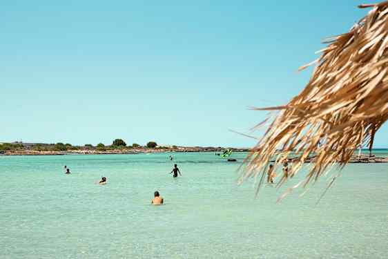 Ranta, jossa on kirkasta vettä ja ihmisiä uimassa ja nauttimassa auringosta. Etualalla näkyy olkikatto. (Tekoälyn luoma teksti)