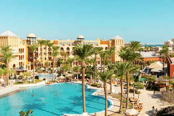 Kuva hotellialueesta, jossa on suuri uima-allas, palmuja ja lämpimän sävyisiä rakennuksia. (Tekoälyn luoma teksti)