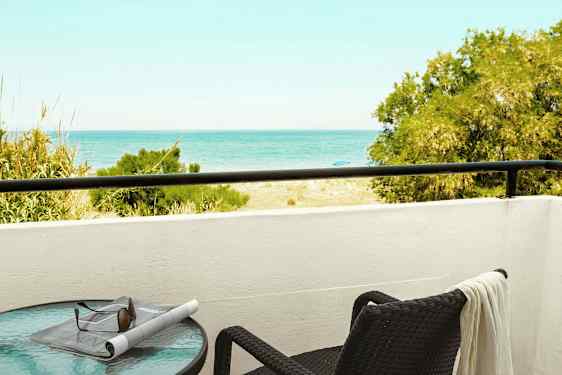 Parveke, josta on näkymä merelle, pöytä aurinkolasien ja sanomalehden kanssa sekä tuoli, jolla on pyyhe. (Tekoälyn luoma teksti)