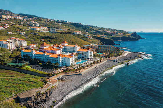 Hotelli, jossa on punaiset tiilikatot meren äärellä, ympäröitynä vihreillä kukkuloilla. (Tekoälyn luoma teksti)