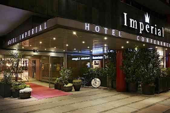 Hotellin ulkokäyttö illalla kyltillä 'Hotel Imperial', ympärillä kasveja ja polkupyörä, jossa kyltti 'Rent Bike'. (Tekoälyn luoma teksti)