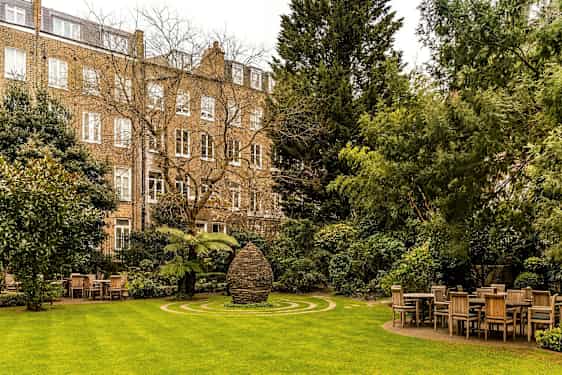 Vihreä puutarha, jossa pöytiä ja tuoleja sekä kivimonumentti keskellä, rakennusten ympäröimänä. (Tekoälyn luoma teksti)