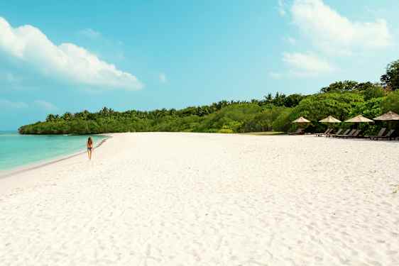 Nainen kävelee hiekkarannalla, jossa taustalla turkoosi vesi ja vihreä kasvillisuus. (Tekoälyn luoma teksti)