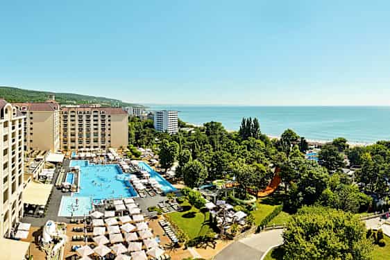 Ilmakuva hotellista, jossa on uima-altaita, aurinkotuoleja ja vehreitä alueita, sijaitsee lähellä merta. (Tekoälyn luoma teksti)