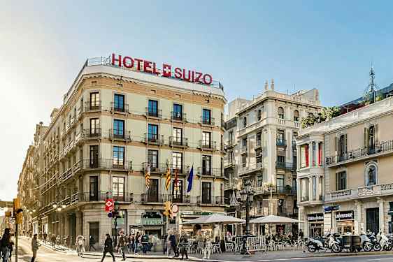 Kadunkulma, jossa on rakennuksia, joista yhdessä lukee hotelli Suizo. Muutamia ihmisiä kulkee ja istuu ulkoterasseilla. (Tekoälyn luoma teksti)