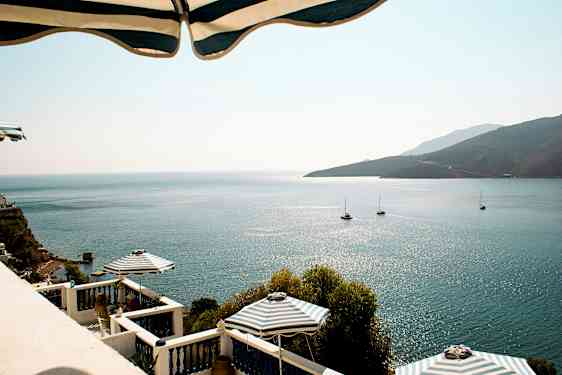 Kuva näkymästä merelle, auringon heijastuksia, purjeveneitä ja terassi aurinkovarjoineen. (Tekoälyn luoma teksti)