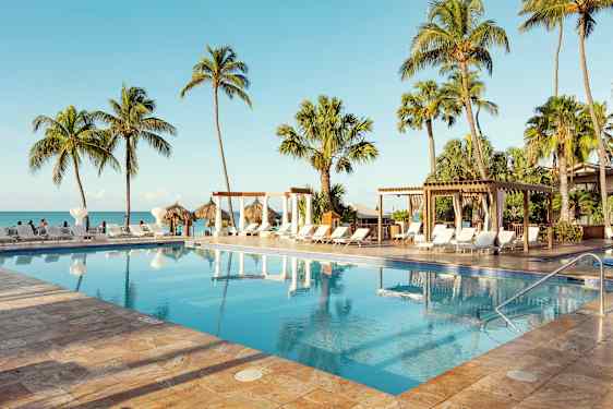 Uima-allas, jota ympäröivät aurinkotuolit, palmut ja pergola, lähellä merta kirkkaassa sinisessä säässä. (Tekoälyn luoma teksti)