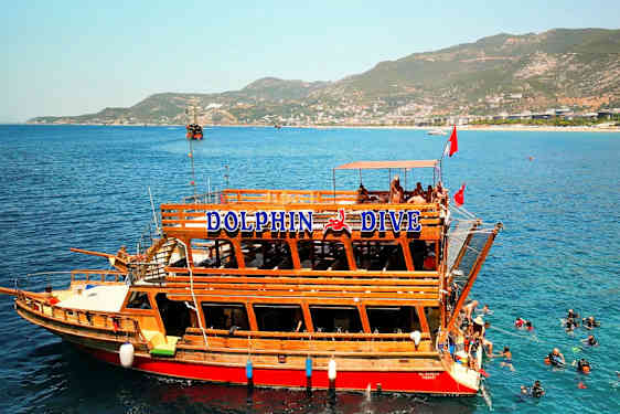 Puisessa veneessä, jossa on teksti "Dolphin Dive", sijaitsee kirkkaansinisen veden äärellä, missä sukeltajat näkyvät vedessä. Taustalla on vuoria ja rantaviiva. (Tekoälyn luoma teksti)