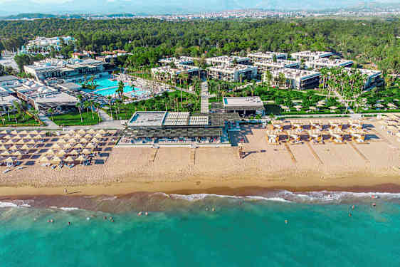 Ilmakuva rantakohteesta, jossa näkyy hiekkaranta, aurinkotuoleja, uima-allasalue ja rakennuksia ympäröivän vehreyden keskellä. (Tekoälyn luoma teksti)