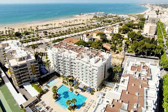 Ilmakuva hotellista, jossa on uima-allasalue, rakennusten ja vehreyden ympäröimänä. Taustalla näkyy hiekkaranta ja meri. (Tekoälyn luoma teksti)