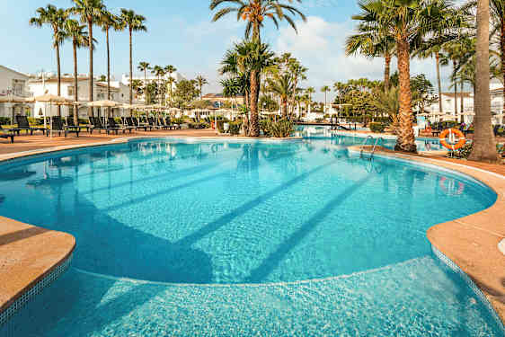 Iso ulkouima-allas, jota ympäröivät aurinkotuolit, aurinkovarjot, palmut ja rakennukset. (Tekoälyn luoma teksti)