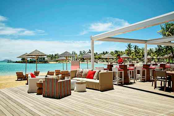 Ulkotarjoilualue rannalla, jossa on baari ja istumapaikkoja, sekä näkymä merelle ja ympäröiviin palmuihin. (Tekoälyn luoma teksti)