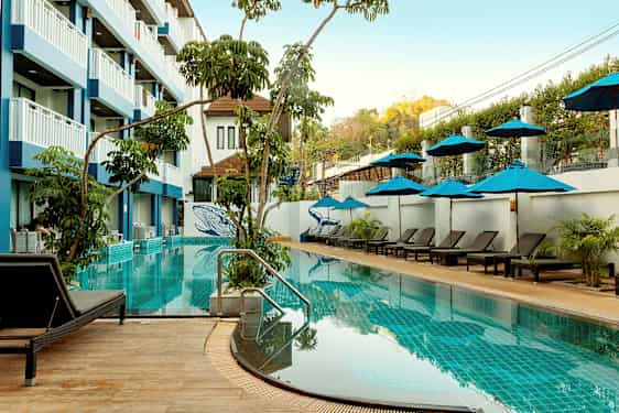 Ulkouima-allas, jossa aurinkotuoleja ja aurinkovarjoja, ympäröitynä kasvillisuudella ja monikerroksisella hotellirakennuksella. (Tekoälyn luoma teksti)