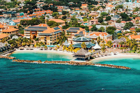 Ilmakuva rannikkokaupungista, jossa turkoosia vettä, hiekkaranta ja rakennuksia punaisilla katoilla ja aurinkopaneeleilla. (Tekoälyn luoma teksti)