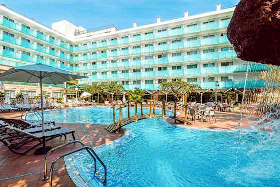 Ulkoallas hotellirakennuksen vieressä, jossa on aurinkotuoleja, pöytiä ja aurinkovarjoja. Pienen sillan yli kulkee osa vesiputousrakennelmaa. (Tekoälyn luoma teksti)