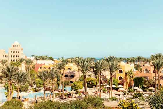 Lomakeskus, jossa on keltaisia ja oransseja rakennuksia, palmuja, vehreää kasvillisuutta sekä uima-altaita ja aurinkovarjoja. (Tekoälyn luoma teksti)