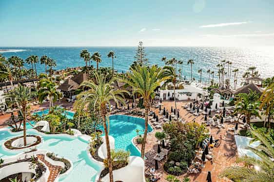 Näköala hotellialueelle, jossa on kaksi uima-allasta, palmuja ja aurinkotuoleja meren rannalla. (Tekoälyn luoma teksti)