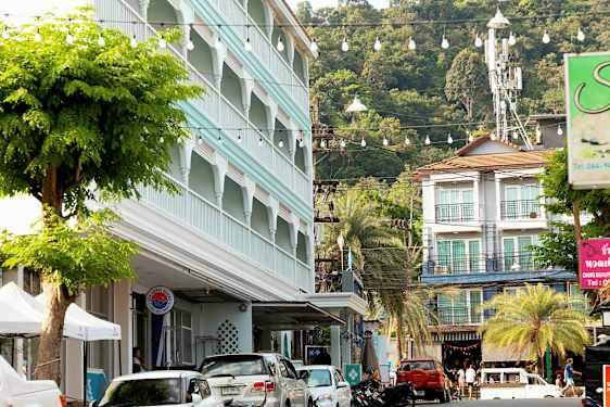 Katu, jossa on pysäköityjä autoja, rakennus sinisellä julkisivulla ja vehmaita puita. (Tekoälyn luoma teksti)