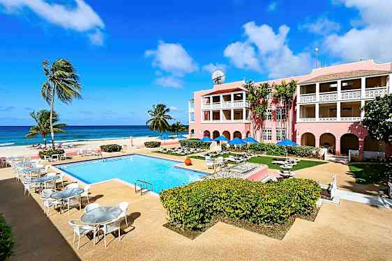 Kuva aurinkoisesta lomakeskuksesta, jossa on ulkouima-allas, ympäröiviä ulkoalueita ja vaaleanpunainen hotelli parvekkeella, meren rannalla. (Tekoälyn luoma teksti)