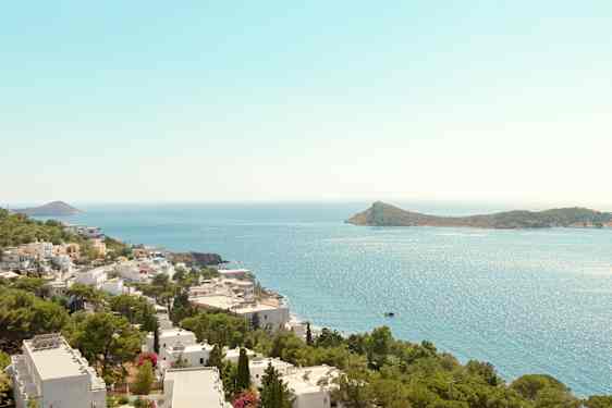 Näkymä rannikon yli, jossa valkoisia rakennuksia ja vihreitä puita, meri ja saari taustalla. (Tekoälyn luoma teksti)