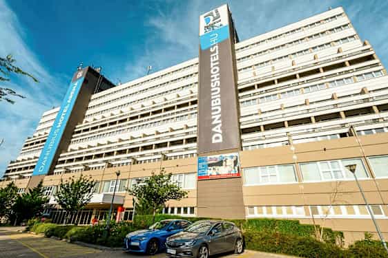 Suuri hotellirakennus, jossa on useita kerroksia ja näkyvässä kyltissä lukee ”Danubius Hotel Arena”. Rakennuksen edessä on pysäköityjä autoja ja joitakin pieniä puita. (Tekoälyn luoma teksti)