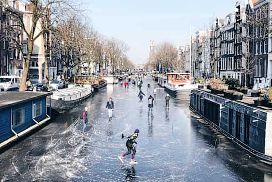 Luisteilijoita ja kävelijöitä jäätyneellä kanavalla, jota asuntolaivat reunustavat.