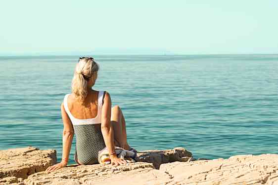 Nainen uimapuvussa istuu rantakivillä ja katsoo merelle.