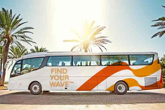 Kuvassa suuri valkoinen lentokenttäbussi, joka on pysäköity palmujen eteen aurinkoisessa lomakohteessa.