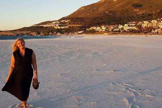 Jutun kirjoittaja kävelee iltavalaistuksessa hiekkarannalla mustassa kesämekossa, sandaalit kädessä.