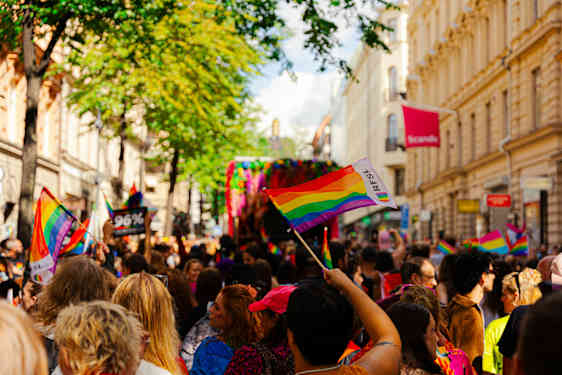 Pride-kulkue täyttää kadun, ja sateenkaariliput liehuvat väkijoukon yllä. Taustalla näkyy värikäs paraativaunu ja kadun varren rakennuksia.