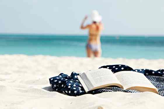 Avoin kirja hiekkarannalla. Taustalla nainen menossa uimaan