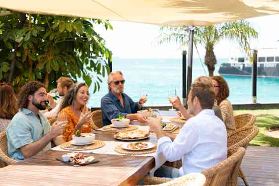 matkaseurue ulkona ruokapöydän ääressä, taustalla näkyy meri.