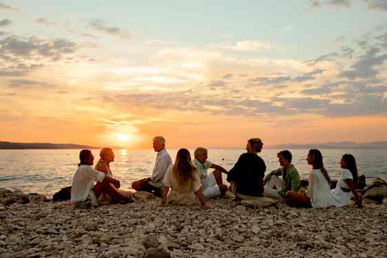 Noin kymmenen hengen porukka istuu pikkukivirannalla auringonlaskussa.