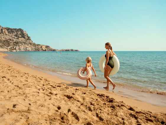 Nainen ja tyttö kävelemässä rannalla uimapuvuissa uimarenkaiden kanssa.