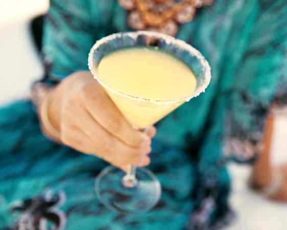 Vaalea cocktail martini-lasissa, taustalla turkoosiin mekkoon pukeutunut henkilö.