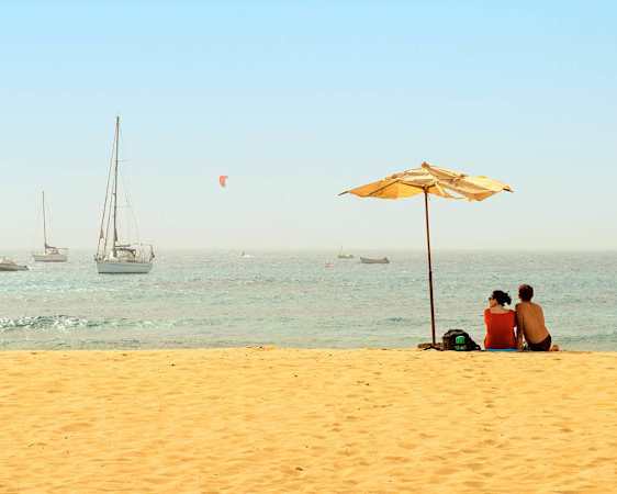 Purjeveneitä merellä ja kaksi henkilöä varjon alla hiekkarannalla