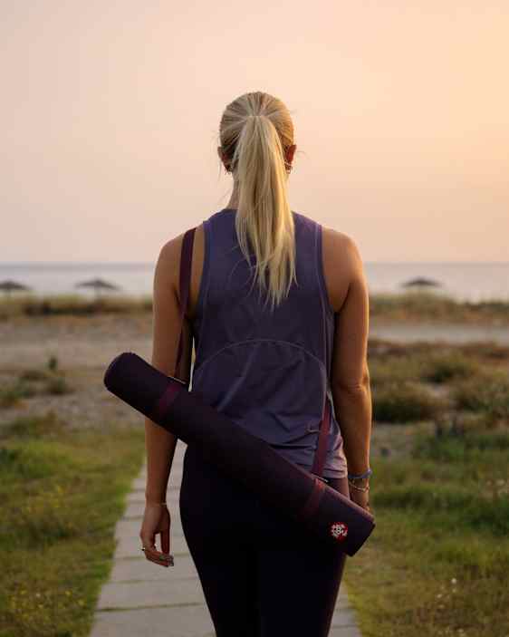 Nainen selin kameraa kävelemässä kohti rantaa joogamatto selässään auringon laskiessa.