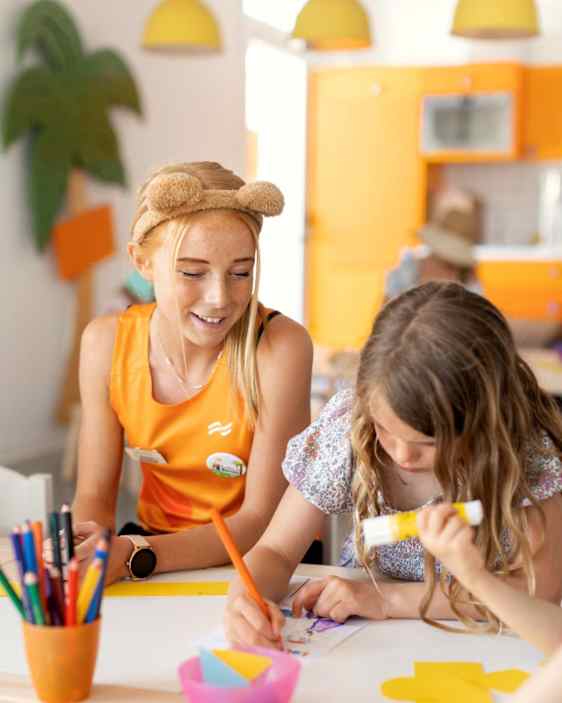 Lastenklubin ohjaaja katsomassa kun tyttö värittää pöydän ääressä.