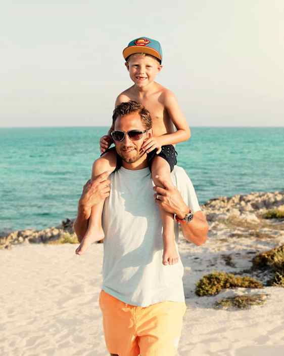 Pieni poika istumassa isänsä harteilla rannalla.