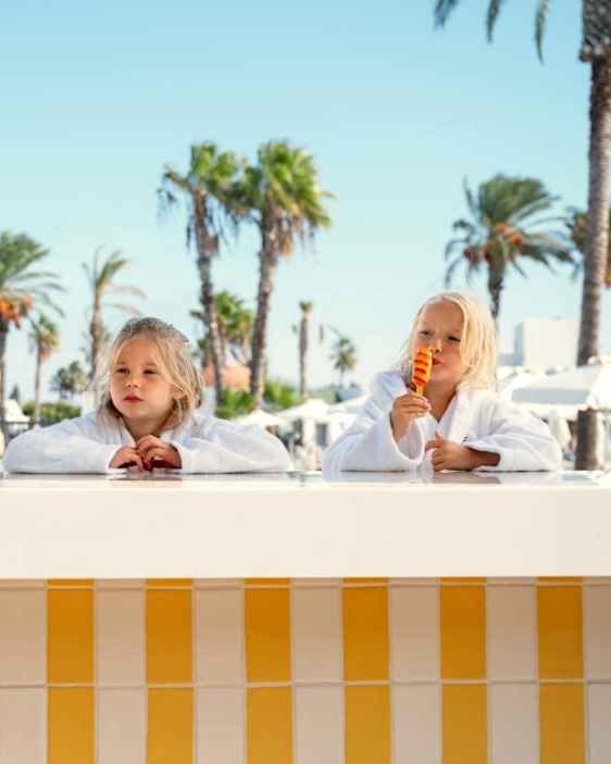 Pienet tytöt allasbaarin tiskillä kypytakeissa, palmupuut ja vesiliukumäki taustalla.