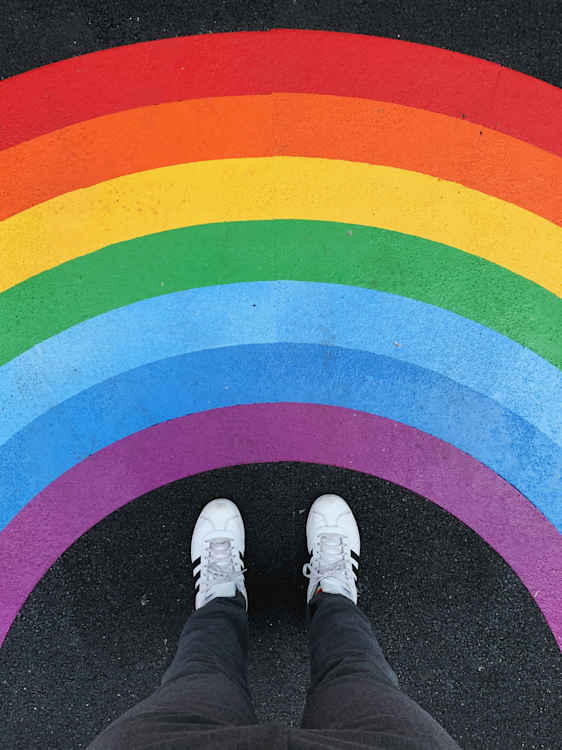 Valkoisiin kenkiin pukeutunut henkilö seisoo maahan maalatun suuren sateenkaarikuvion päällä.