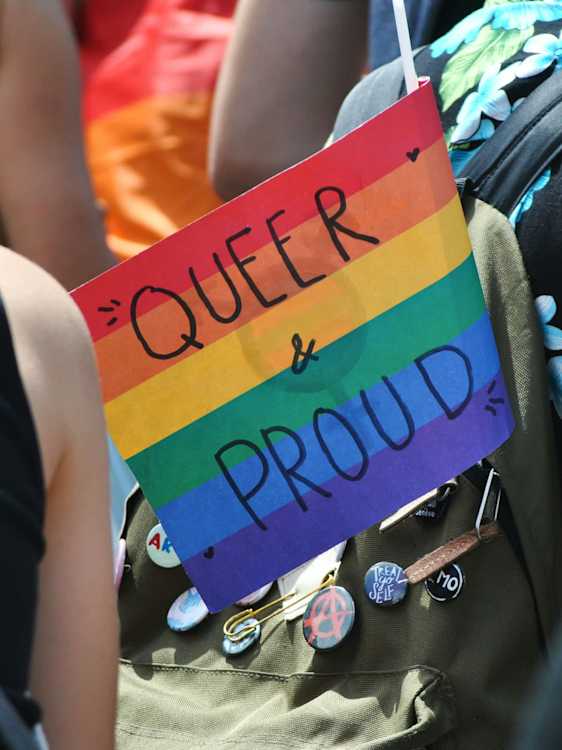 Kädessä pideltävä sateenkaarilippu, jossa lukee ”Queer & Proud”, Pride-tapahtuman ihmisvilinässä.