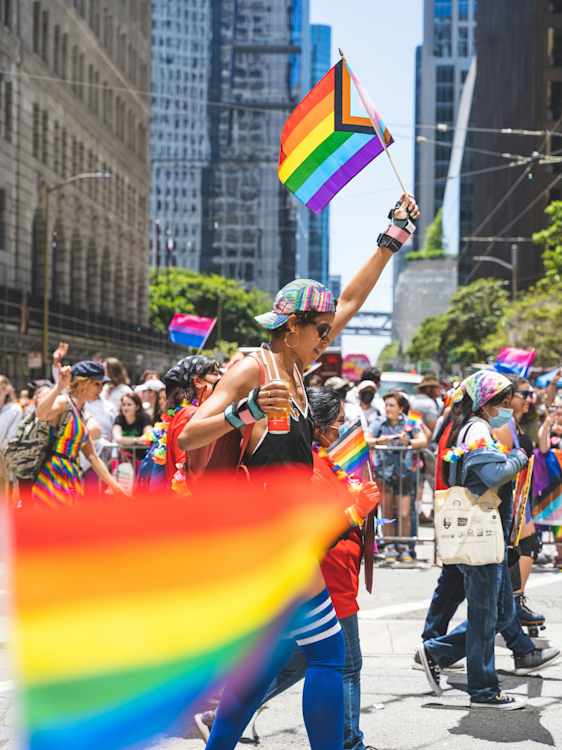 Pride-kulkue täyttää kadun ihmisillä, sateenkaarilipuilla ja värikkäillä asuilla. Taustalla kohoavat kaupungin pilvenpiirtäjät.