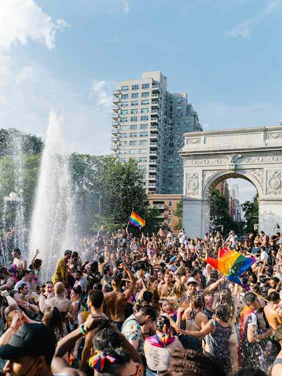 Suuri väkijoukko juhlii Pridea puistossa. Sateenkaariliput liehuvat osallistujien keskellä, ja taustalla näkyy riemukaari ja korkeita rakennuksia.