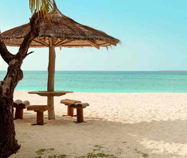 Aurinkovarjo, puinen pöytä ja istumapaikat hiekkarannalla, meri taustalla. (Tekoälyn luoma teksti)