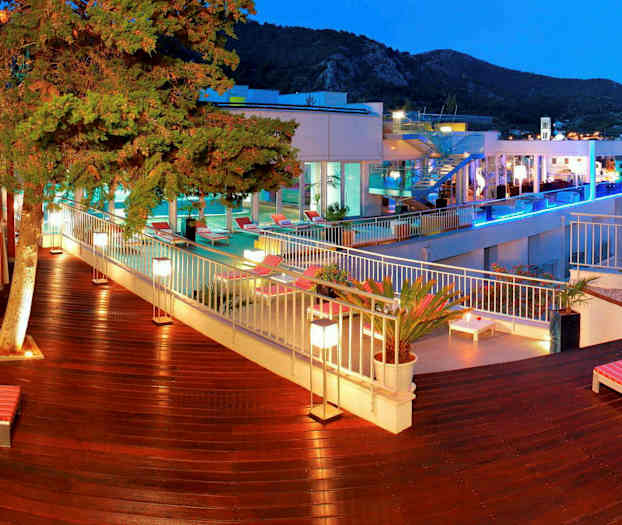 Ulkotila, jossa on puuterassi, aurinkotuoleja ja vihreitä kasveja uima-altaan läheisyydessä. Taustalla näkyy rakennuksia ja vuoristomaisema. (Tekoälyn luoma teksti)