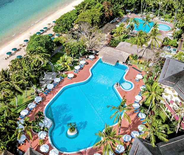 Ilmakuva uima-allasalueesta, jota ympäröivät trooppiset puut, ranta ja meri taustalla. (Tekoälyn luoma teksti)